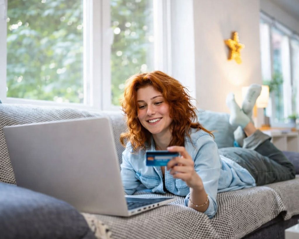 Frau liegt mit Laptop und Kreditkarte auf dem Sofa und kauft online ein